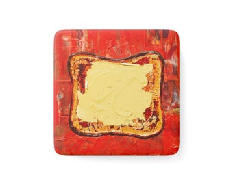 May include: A square coaster featuring a painted image of a slice of toast topped with a thick layer of yellow butter. The background is a vibrant, textured red with visible brushstrokes, and the toast has a dark brown crust and golden centre.