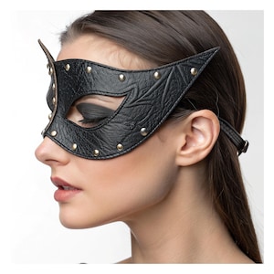 Könnte beinhalten: Schwarze Leder-Katzenaugenmaske mit goldfarbenen Nieten. Die Maske hat eine strukturierte Oberfläche und wird mit einem dünnen Riemen befestigt. Die Maske bedeckt die Augen und hat ein spitzes Ohrendesign.
