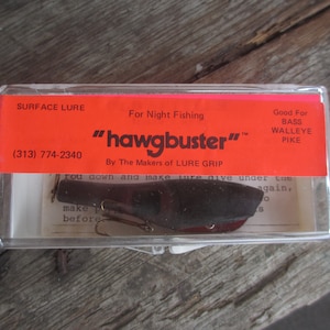 May include: A red and white plastic container with a black fishing lure inside. The container has the text "SURFACE LURE" and "hawgbuster" on it. The lure is designed for night fishing and is good for bass, walleye, and pike.