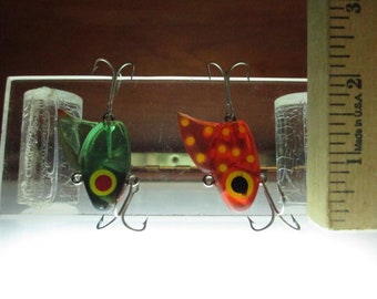 May include: Two fishing lures, one green with red and yellow accents and one red with yellow and black accents. The lures are displayed on a clear surface with a ruler in the background.