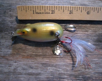 May include: A vintage fishing lure shaped like a frog, with a yellow and black body, silver spinner blades, and white feathers. The lure is approximately 4 inches long, with a wooden ruler in the background.