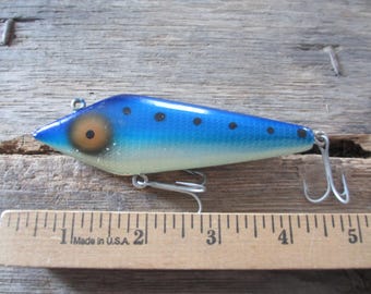 May include: A blue and white fishing lure with black dots and a large eye design. The lure has two treble hooks and is approximately 4 inches long. The lure is lying on a wooden surface.