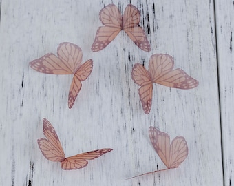 May include: Six delicate, translucent butterfly decorations in a light peach color, scattered on a white wooden surface.