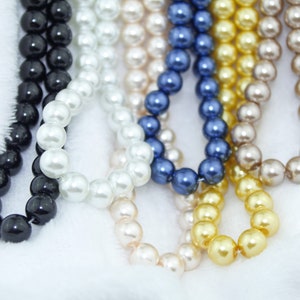 May include: A collection of six pearl necklaces in various colors, including black, white, cream, light blue, gold, and brown. The necklaces are made of round pearls and are arranged in a layered fashion.
