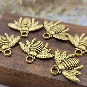 May include: A collection of gold-toned bee charms with intricate details. The charms are arranged on a wooden surface and feature a variety of poses, including flying, resting, and crawling.