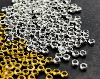 May include: A close-up of a collection of small, metallic beads. The beads are scattered across a dark surface, with a mix of gold and silver colours. The beads are round with a hole through the centre, suitable for jewellery making or crafts.