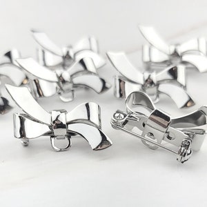 May include: A set of silver-toned bow-shaped brooches with a pin fastening. The brooches are arranged in a cluster on a white surface.