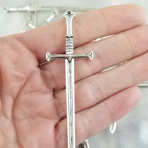 May include: Silver-toned sword charm with a cross guard and a pointed tip. The sword is about 2 inches long.