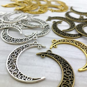 Puede incluir: Una colección de dijes de media luna de filigrana plateada y dorada. Los dijes son intrincadamente detallados y tienen un diseño delicado y ornamentado.