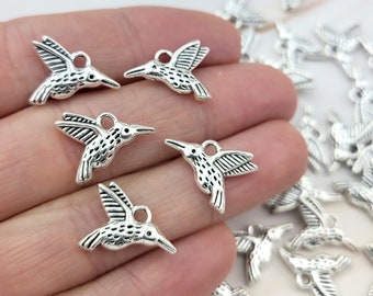May include: A hand holding a group of silver hummingbird charms. The charms are detailed and have a vintage look.