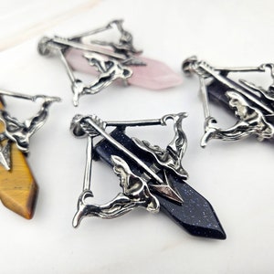 May include: Three silver arrow pendants with different colored gemstones. The pendants feature a detailed design of an arrow and a figure holding the arrow. The gemstones are pink, purple, and blue.
