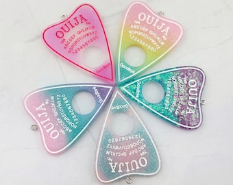 Puede incluir: Cuatro encantos de planchette Ouija de resina, llenos de purpurina, con forma de corazón y un agujero en la parte superior para colgar. Cada encanto tiene un degradado de color diferente y presenta las palabras "Ouija" y las letras del alfabeto, los números del 1 al 9 y el 0, así como las palabras "Sí", "No" y "Adiós".