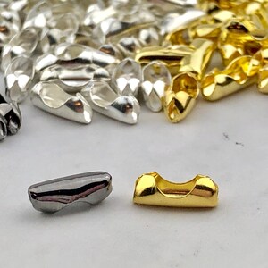 Puede incluir: Primer plano de varios hallazgos de cuentas de crimpado met&aacute;licas en colores plata, oro y metal de ca&ntilde;&oacute;n. Las cuentas son peque&ntilde;as, oblongas y tienen un dise&ntilde;o curvo. Estos componentes para la fabricaci&oacute;n de joyas se utilizan para asegurar cierres.
