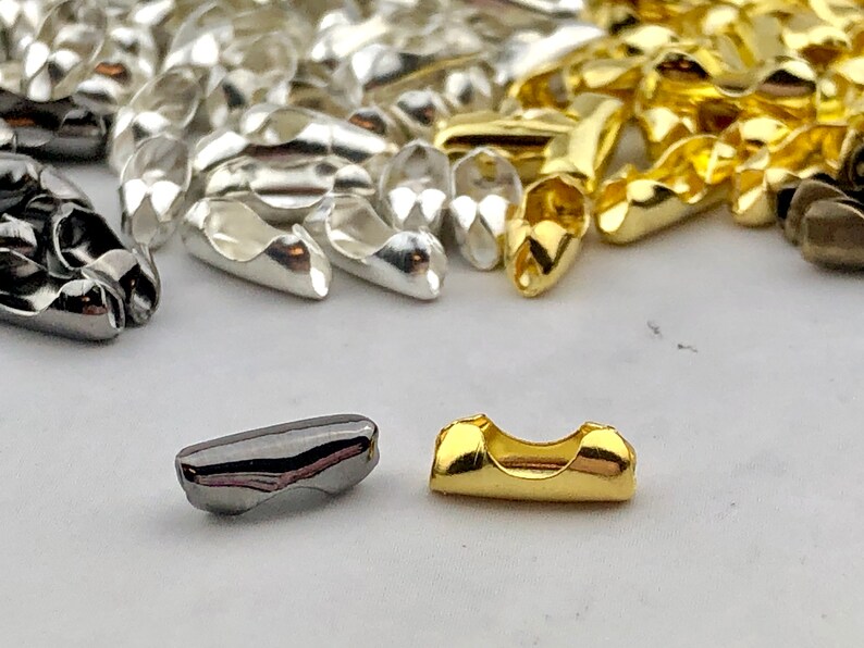 Puede incluir: Primer plano de varios hallazgos de cuentas de crimpado met&aacute;licas en colores plata, oro y metal de ca&ntilde;&oacute;n. Las cuentas son peque&ntilde;as, oblongas y tienen un dise&ntilde;o curvo. Estos componentes para la fabricaci&oacute;n de joyas se utilizan para asegurar cierres.