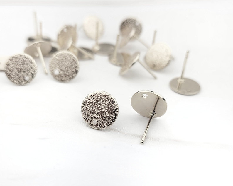 May include: Silver earring blanks with a textured round surface and a hole in the center. The earring posts are attached to the back of the round surface.