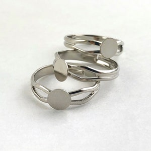 Puede incluir: Tres anillos plateados con un diseño minimalista. Cada anillo presenta una pieza central circular y plana y una banda dividida. Los anillos se exhiben sobre una superficie blanca, mostrando su elegancia simple y estilo moderno.