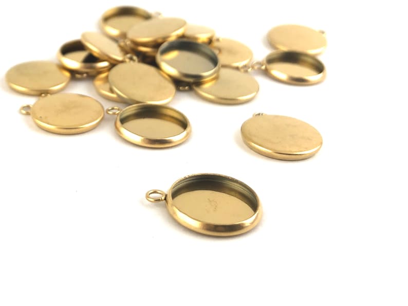 May include: A group of gold-toned metal pendant trays with a round shape and a smooth surface. The trays have a small loop at the top for attaching to a chain or cord.