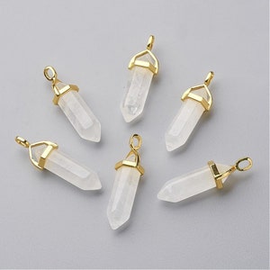 May include: Six clear quartz crystal pendants with gold-colored metal caps and loops. The crystals are hexagonal in shape and have a polished, translucent appearance. The pendants are arranged on a light gray surface.