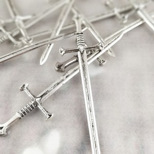 May include: A group of silver toy swords, some with intricate details, are scattered on a white surface. The swords are arranged in a random pattern, with some overlapping each other.