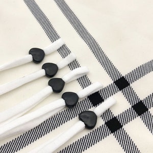 Puede incluir: Cinco cordones elásticos blancos con tapones de plástico en forma de corazón negro. Los cordones están dispuestos en forma de abanico sobre una superficie a cuadros blancos y negros.