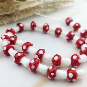 Puede incluir: Primer plano de cuentas en forma de hongo. Las cuentas tienen tapas rojas con lunares blancos y tallos blancos. Las cuentas están ensartadas en un hilo transparente. El fondo es blanco suave.