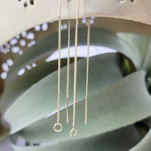 May include: A pair of gold chain earrings with small circular charms at the bottom. The earrings are hanging from a white surface with a decorative pattern.