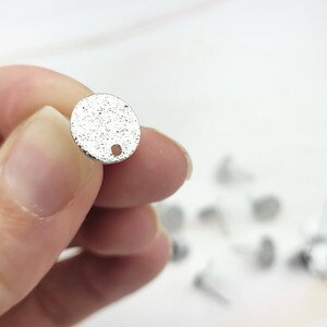 May include: Silver round metal charm with a textured surface and a small hole at the top.