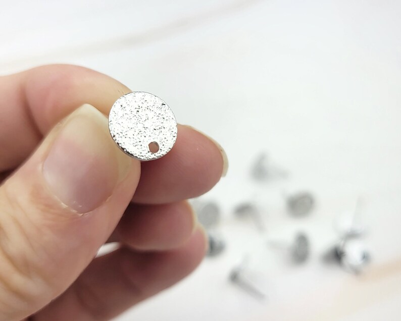 May include: Silver round metal charm with a textured surface and a small hole at the top.