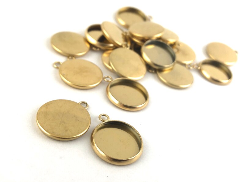 May include: A group of gold-toned metal round pendant trays with a smooth, shiny finish. The trays have a lip around the edge and a small loop at the top for attaching to a chain or cord.