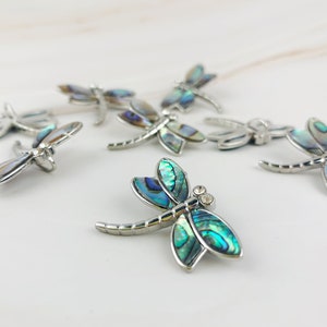 May include: A group of six silver dragonfly charms with iridescent blue and green abalone shell wings. The charms have a silver finish and are scattered on a white marble surface.