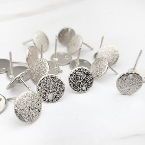 May include: A close-up of a pile of silver earring blanks. The blanks are round and have a textured surface. They are designed to be used with a variety of materials, such as beads, stones, and charms.