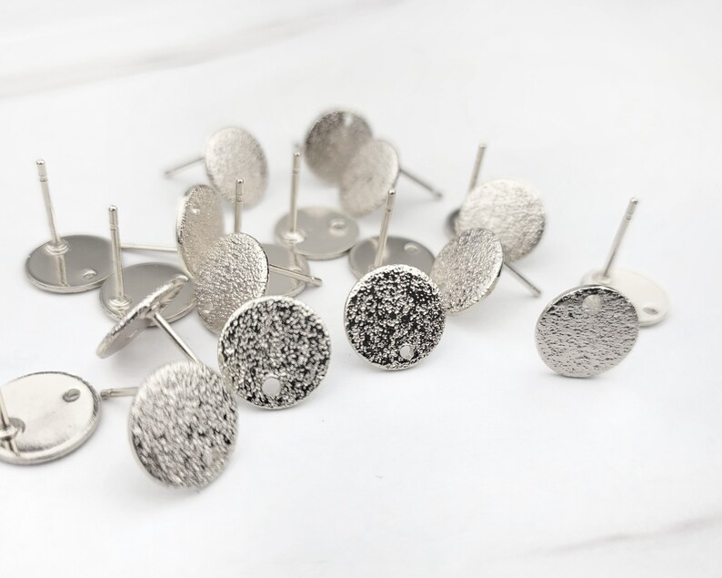 May include: A close-up of a pile of silver earring blanks. The blanks are round and have a textured surface. They are designed to be used with a variety of materials, such as beads, stones, and charms.