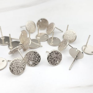 May include: A group of silver-toned earring blanks with a textured surface. The earring blanks are round and have a small hole in the center.
