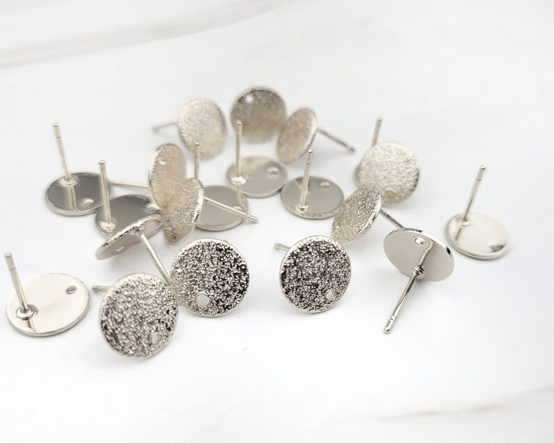 May include: A group of silver-toned earring blanks with a textured surface. The earring blanks are round and have a small hole in the center.