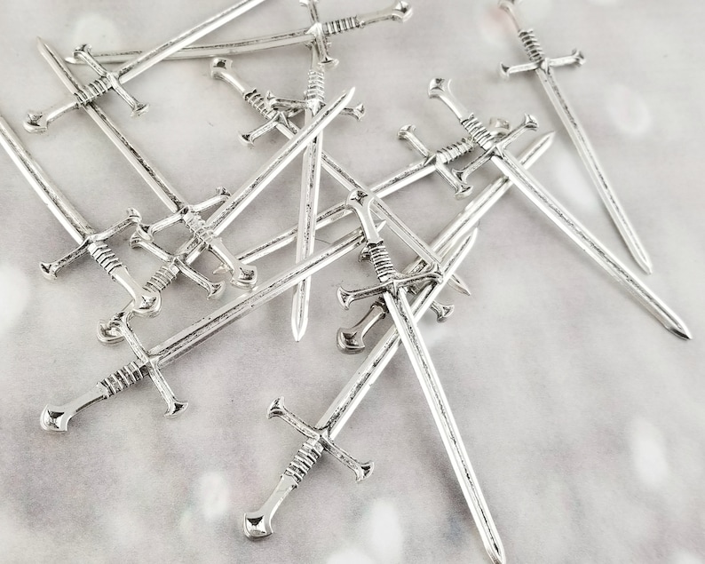 May include: A group of silver-toned sword-shaped cocktail picks. The picks are arranged in a random pattern, with some overlapping.
