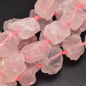 May include: A strand of pink rose quartz beads, each bead is a natural, rough-cut, irregular shape. The beads are strung on a pink thread.