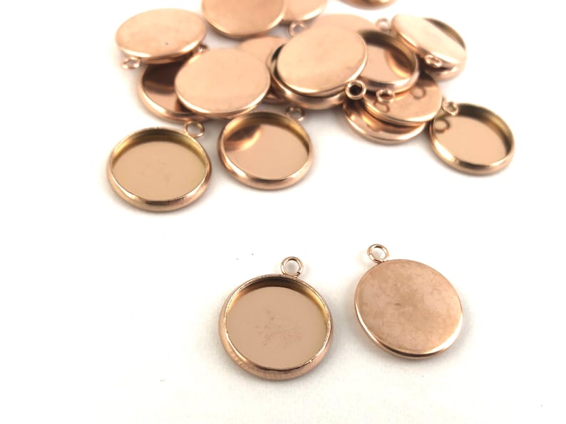 May include: A group of rose gold metal round pendant trays with a loop for hanging. The trays are empty and ready to be filled with resin, epoxy, or other materials.