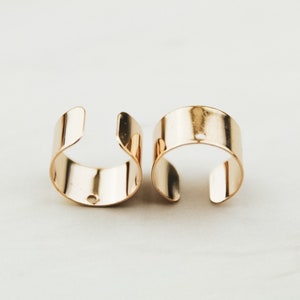 May include: Two gold-toned metal ear cuffs. Each cuff is a simple, rounded band with a small hole in the center.