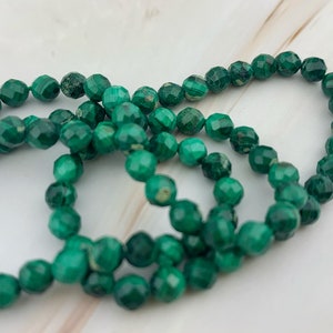 May include: A strand of green malachite beads, each bead is faceted and has a unique pattern. The beads are strung together on a clear thread.