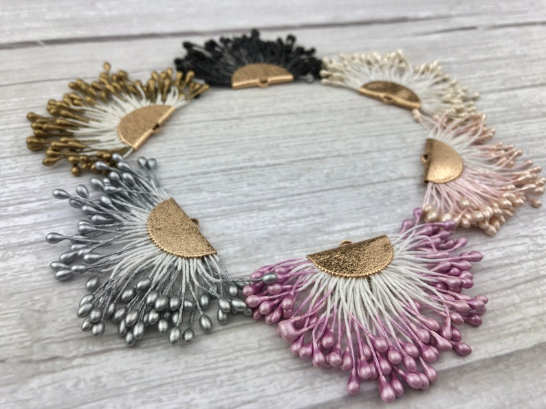 May include: Six decorative fan-shaped embellishments with a gold metal accent. The embellishments are in various colors, including black, white, gold, silver, and pink.