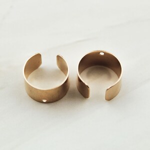 May include: Two gold-colored metal rings with a small hole in each. The rings are open and have a rounded shape.