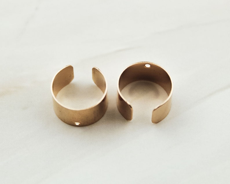 May include: Two gold-colored metal rings with a small hole in each. The rings are open and have a rounded shape.