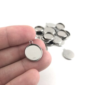 May include: A close-up of a silver pendant with a round, clear glass insert. The pendant is being held by a hand. There are several other similar pendants in the background.