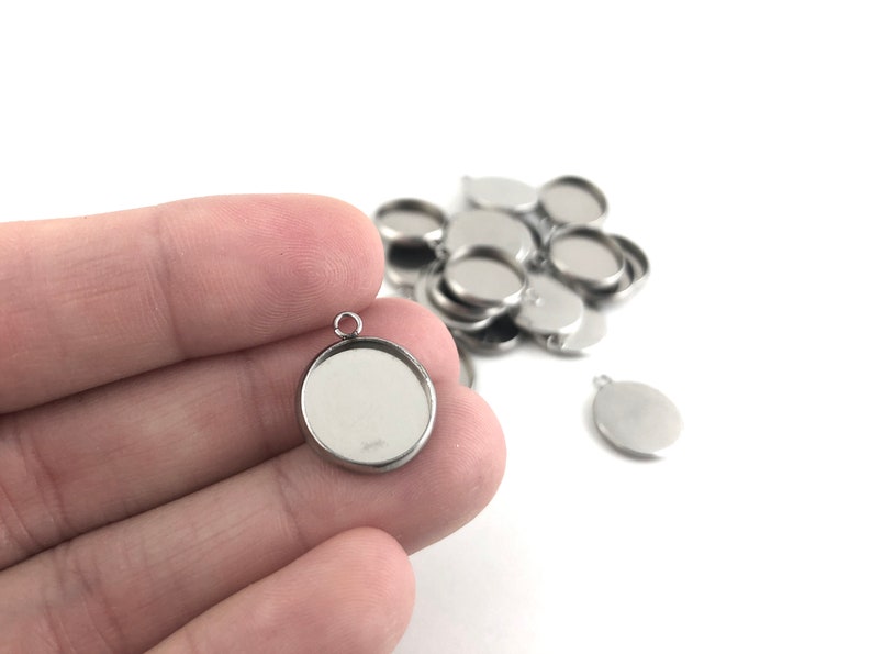 May include: A close-up of a silver pendant with a round, clear glass insert. The pendant is being held by a hand. There are several other similar pendants in the background.