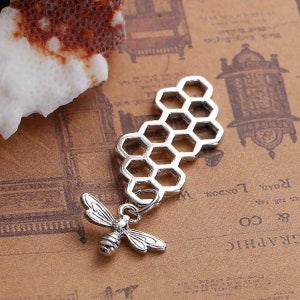 May include: Silver charm featuring a honeycomb design with a small bee pendant. The charm is made of metal and has a shiny finish.