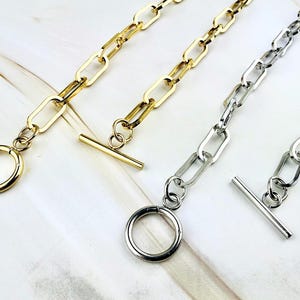 Könnte beinhalten: Nahaufnahme von goldenen und silbernen Kettenhalsketten mit Knebelverschlüssen. Die Halsketten haben längliche ovale Glieder und einen kreisförmigen Ring mit einem Stangenverschluss. Der Schmuck wird auf einer weißen Oberfläche präsentiert.