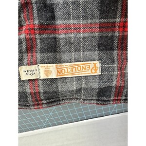 May include: A grey, black, and red plaid wool fabric featuring a rectangular white tag with the text Pendleton, 100% Virgin Wool, Portland, Oregon, and dry clean. The fabric has a woven texture with a repeating pattern of intersecting lines.