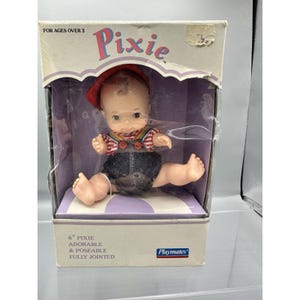 May include: A vintage Playmates Pixie doll in its original box. The doll is 6 inches tall and fully jointed, wearing a red cap, a striped shirt, denim overalls, and rainbow suspenders. The box reads "For Ages Over 3".