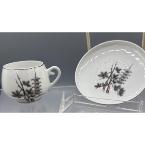 May include: White teacup and saucer set with a Japanese pagoda and tree design. The cup has a curved handle and a silver rim. The saucer is round and also features the same pagoda and tree design. The set is on a clear display stand.