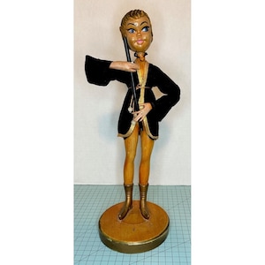 May include: A vintage wooden doll with a painted face, wearing a black velvet jacket with gold trim and matching boots. The doll is holding a black rod and stands on a round wooden base.
